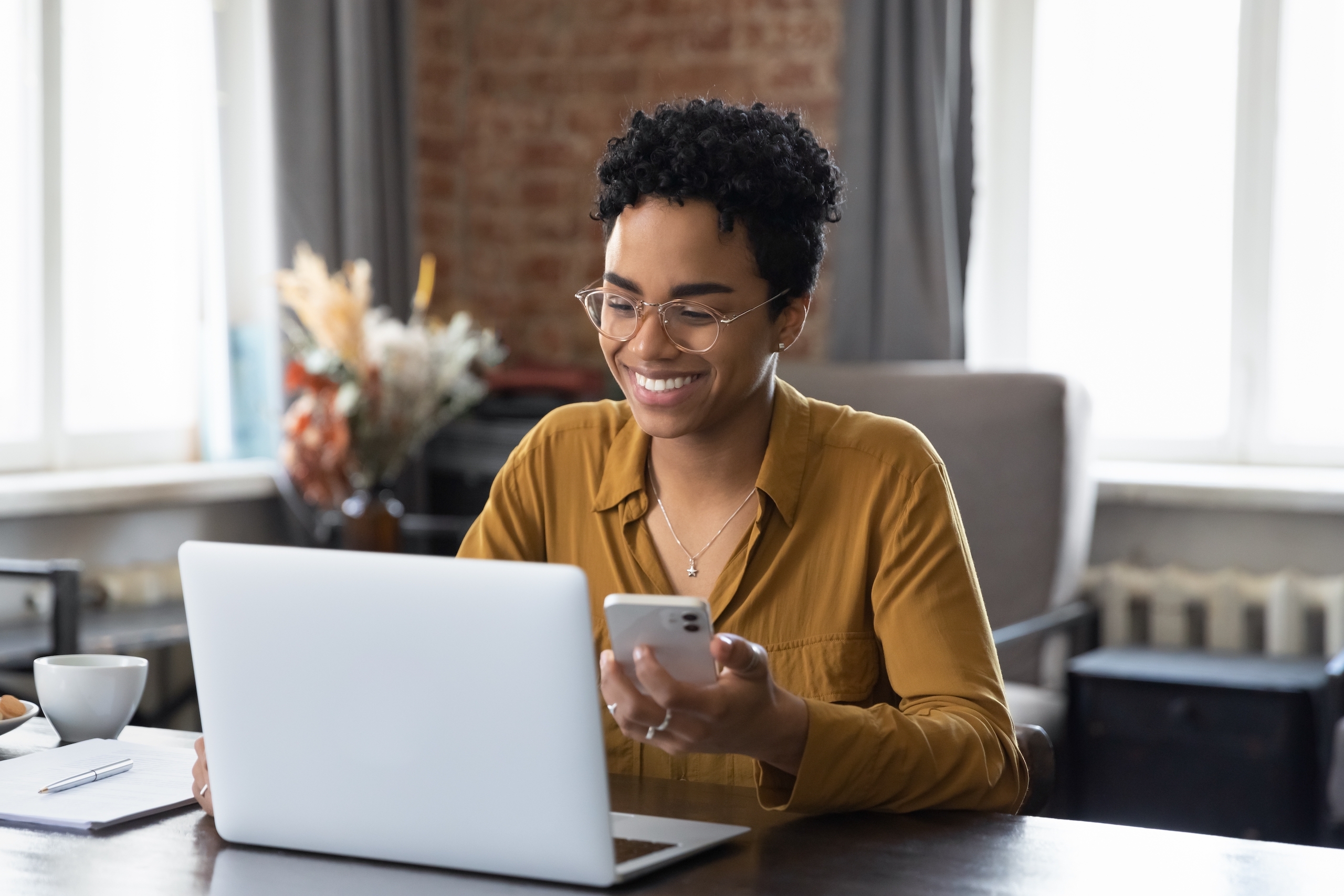 A woman happily multitasks, smiling as she uses her laptop and cell phone simultaneously.