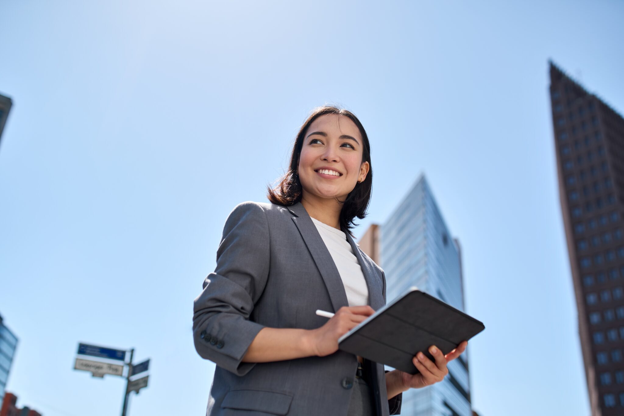 An Asian businesswoman confidently holds a tablet while standing in a bustling city.